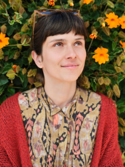 Una mujer de pelo castaño corto y flequillo, vestida con una camisa estampada y una rebeca roja, descansa sobre un follaje verde con vibrantes flores naranjas, mirando hacia arriba con una amable sonrisa. Sobre su cabeza reposan unas gafas de sol.