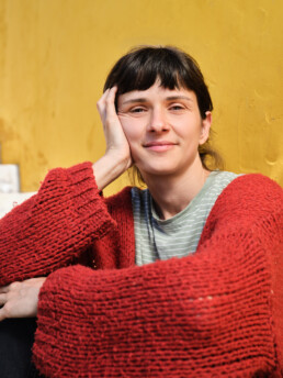 Una persona de pelo y flequillo oscuros, con un jersey de punto rojo y una camisa de rayas, se sienta contra una pared amarilla, apoya la cara en una mano y sonríe suavemente a la cámara.