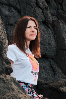 Una mujer de pelo largo y castaño está de pie entre rocas volcánicas oscuras, con una camiseta blanca de Hawai con un gráfico de una puesta de sol rosa y amarilla y una falda de flores. Mira al frente con una leve sonrisa.