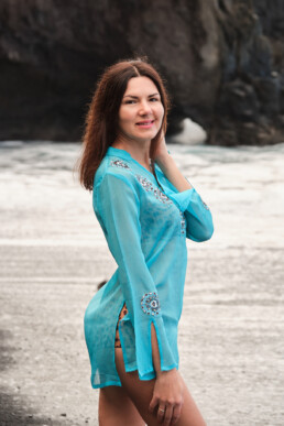 Una mujer de pelo largo y castaño, vestida con un encubrimiento azul, está de pie en una playa, sonriendo a la cámara. Al fondo se ven acantilados rocosos y arena.