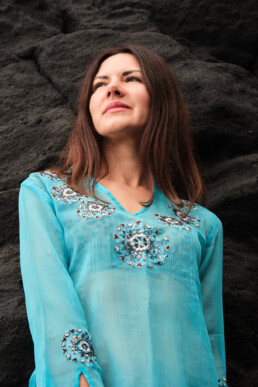 Una mujer de pelo largo y castaño, con un top bordado azul claro, está de pie delante de un fondo de roca oscura y áspera, mirando ligeramente hacia un lado con una sonrisa amable.